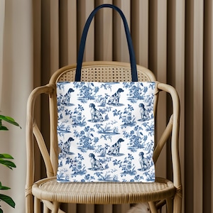 Può includere: Una borsa tote bianca con un motivo toile de Jouy blu con cani ed elementi floreali. La borsa ha un manico blu navy ed è esposta su una sedia di vimini. Il design della borsa ricorda i classici tessuti toile.