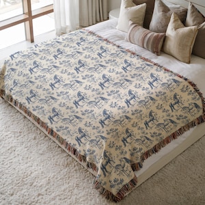 May include: A cream-coloured throw blanket featuring a blue horse toile pattern. The blanket has a fringed edge with red, brown, and blue threads. The blanket is on a bed with several pillows.