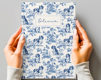 Puede incluir: Una libreta blanca con un patrón azul y blanco de perros y flores. El nombre "Olivia" está impreso en la portada de la libreta.