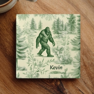 Könnte beinhalten: Quadratischer Untersetzer mit einer grünen Bigfoot-Illustration, die durch eine Waldszene geht. Der Hintergrund zeigt ein sich wiederholendes Muster aus Bäumen und Blättern in Grüntönen. Der Name "Kevin" ist in einer Schriftart gedruckt.