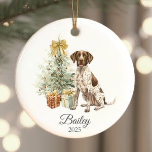 May include: White ceramic Christmas ornament featuring a brown and white dog, a decorated Christmas tree, and wrapped gifts. The ornament has a gold ribbon for hanging and includes the name "Bailey" and the year "2025".