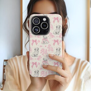 Könnte beinhalten: Eine weiße iPhone-Hülle mit einem rosa und grauen Muster von Hunden, die Schleifen tragen. Die Hülle hat einen Ausschnitt für das Kameraobjektiv und einen Ausschnitt für die Tasten des Telefons.