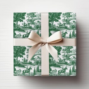May include: A wrapped gift box featuring a green and white toile pattern with riders on horseback, trees, and bridges. The box is tied with a beige ribbon and bow, creating a classic and elegant presentation.