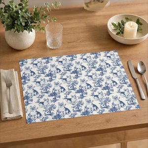Puede incluir: Un mantel individual rectangular con un estampado de toile de Jouy azul y blanco con perros Corgi y follaje. El mantel individual está sobre una mesa de madera, junto a una servilleta, cubiertos y un vaso.
