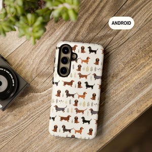 May include: A white phone case with a pattern of brown and black dachshund dogs on a white background. The case has a camera cutout and the word "ANDROID" is printed on the top right corner.