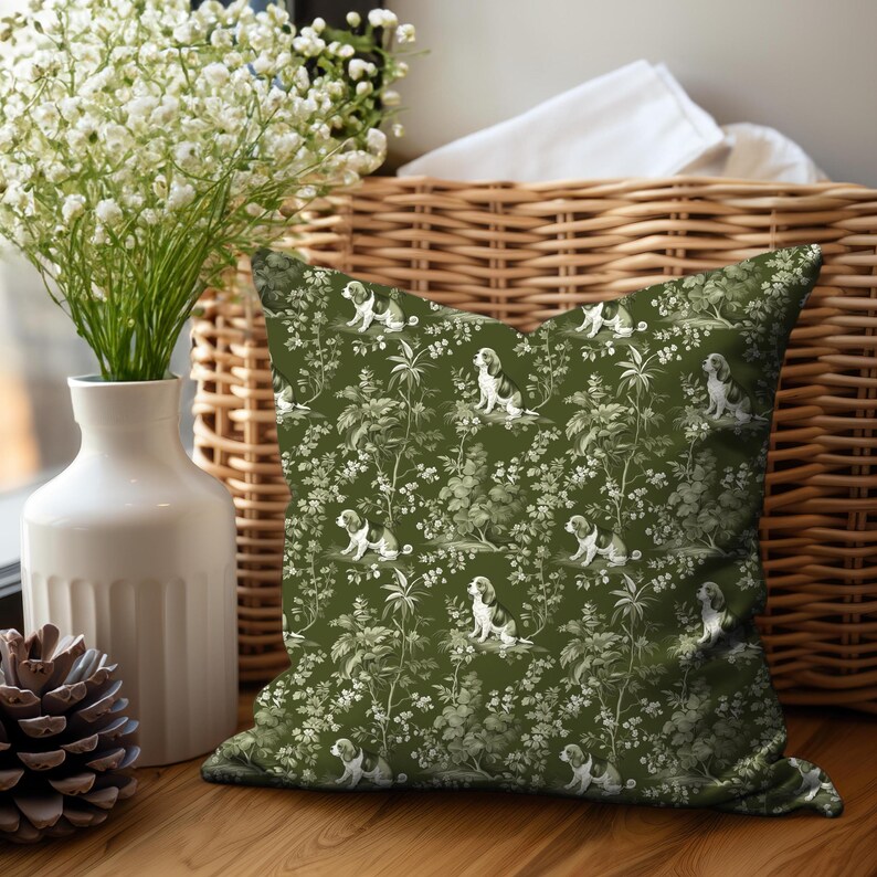 May include: A green throw pillow with a white and gray pattern of dogs and foliage. The pillow is sitting on a wicker basket.