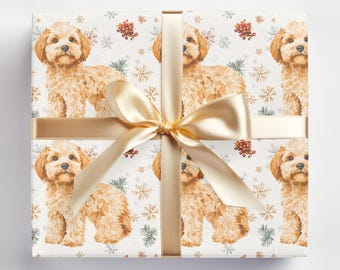 Puede incluir: Papel de regalo blanco con un patrón repetido de perros marrones y blancos, copos de nieve y bayas rojas. Una cinta dorada está atada alrededor de la caja con un lazo.
