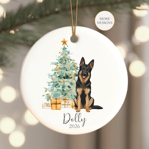 May include: A round white ceramic ornament featuring a watercolor illustration of a German Shepherd sitting next to a decorated Christmas tree with gold gifts underneath. The text "Dolly" and "2026" is printed below the dog. A gold string is attached.