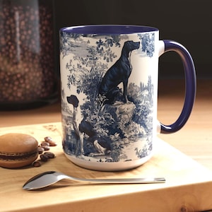 Puede incluir: Taza de cerámica blanca con un diseño toile de Jouy azul y blanco con dos perros en un paisaje. La taza tiene un asa y un borde azul oscuro. Una cuchara y un macaron están sobre una superficie de madera.