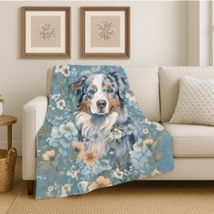 May include: A soft throw blanket featuring a blue merle Australian Shepherd dog portrait surrounded by a pattern of light blue, white, and soft orange flowers. The blanket has a muted color palette and a detailed illustration of the dog.