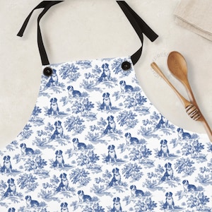 May include: A white apron with a blue and white toile pattern featuring dogs. The apron has adjustable straps and a pocket.