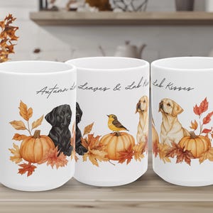 May include: Three white ceramic mugs featuring a repeating autumn theme. The design includes illustrations of black and yellow Labrador dogs, orange pumpkins, and fall leaves. The text "Autumn Leaves & Lab Kisses" is printed in black script.