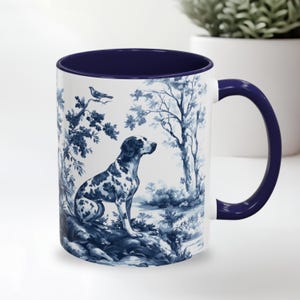 Puede incluir: Una taza de cerámica blanca con un diseño azul y blanco de un perro en un entorno de bosque. La taza tiene un borde y un asa azules.
