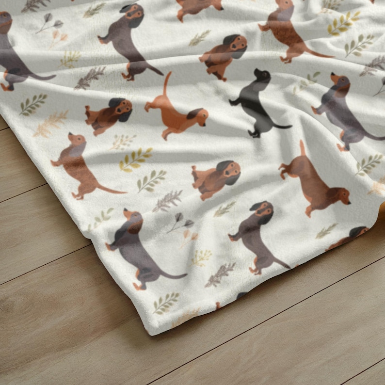 May include: A white blanket with a repeating pattern of dachshund dogs in various shades of brown and black. The blanket also features small green and yellow floral accents, creating a playful and cozy design.