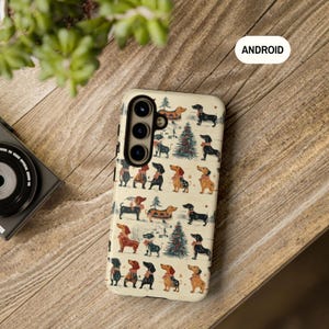 May include: A white phone case with a pattern of dachshunds wearing Christmas sweaters and hats. The case has a black border and a camera cutout. The text "ANDROID" is printed on the case.
