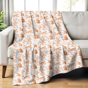 May include: A white throw blanket draped over a sofa, featuring a repeating pattern of orange cats and trees. The blanket's design resembles a toile print, adding a touch of classic style to the room. The blanket is made of a soft material.