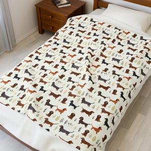 May include: A cream-colored blanket featuring a repeating pattern of dachshund dogs in various colors, including brown, black, and gray. The blanket also has small leaf accents. The blanket is spread over a bed.