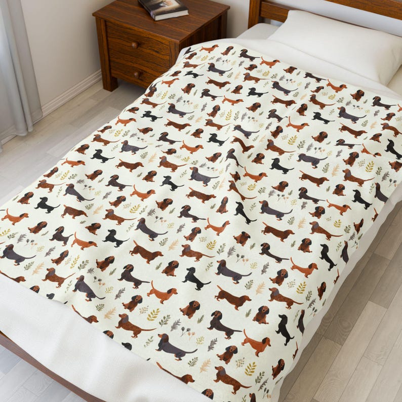 May include: A cream-colored blanket featuring a repeating pattern of dachshund dogs in various colors, including brown, black, and gray. The blanket also has small leaf accents. The blanket is spread over a bed.