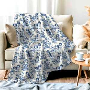 Può includere: Una coperta bianca e blu con un motivo ripetuto di cani in un design toile. I cani sono in posizione seduta e sono circondati da motivi floreali.