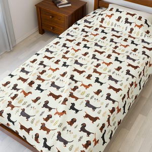 May include: A cream-colored twin-size bedspread featuring a repeating pattern of dachshund dogs in various shades of brown and black. The design includes small green and brown leaf accents, creating a playful and whimsical aesthetic.