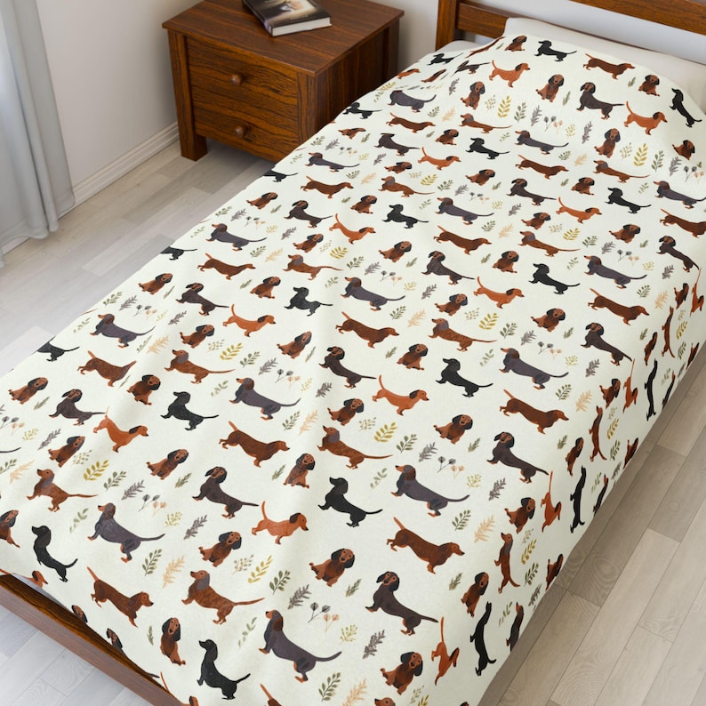 May include: A cream-colored twin-size bedspread featuring a repeating pattern of dachshund dogs in various shades of brown and black. The design includes small green and brown leaf accents, creating a playful and whimsical aesthetic.