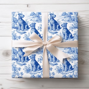 May include: A wrapped gift with a cream-colored satin bow. The wrapping paper features a blue and white toile pattern with illustrations of dogs, trees, and landscapes. The gift is presented on a white wooden surface.