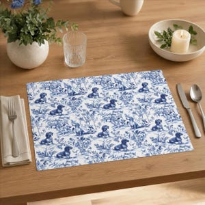 Può includere: Un sottopiatto rettangolare con un motivo toile de Jouy blu e bianco con cani bassotto in un ambiente paesaggistico. Il sottopiatto è su un tavolo di legno, con una forchetta, un coltello e un cucchiaio a destra.