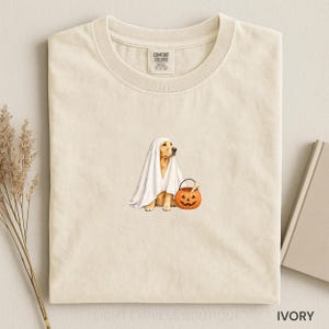 May include: An ivory-colored crew neck t-shirt featuring a graphic of a golden retriever wearing a white ghost costume and holding an orange jack-o'-lantern pumpkin bucket. The shirt has a Comfort Colors brand label on the inside neck area.