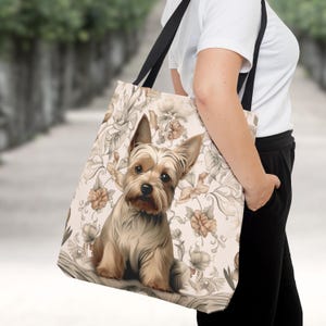 Pu&ograve; includere: Una borsa tote con un'illustrazione di un Yorkshire Terrier color marrone e beige centrata su uno sfondo beige chiaro con un motivo ripetitivo di fiori dai toni tenui e linee curve, dotata di due tracolle nere.