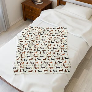 May include: A cream-colored blanket featuring a repeating pattern of dachshund dogs in various colors, including brown, black, and tan. The blanket is placed on a white bed, creating a cozy and inviting aesthetic.