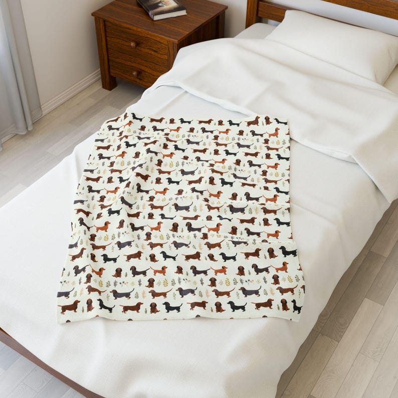 May include: A cream-colored blanket featuring a repeating pattern of dachshund dogs in various colors, including brown, black, and tan. The blanket is placed on a white bed, creating a cozy and inviting aesthetic.