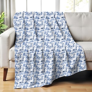 May include: A white and blue throw blanket draped over a white sofa. The blanket features a repeating pattern of a dog in a landscape scene. The color scheme is reminiscent of classic blue and white porcelain.