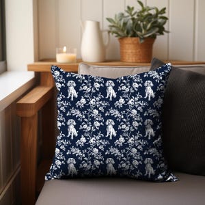 May include: A square decorative pillow with a navy blue background and a white floral and dog pattern. The pillow is placed on a gray cushion, with a wooden armrest and a window in the background.