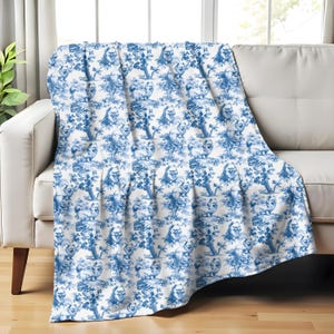 Könnte beinhalten: Eine blau-weiße Decke, die über einem weißen Sofa drapiert ist. Die Decke zeigt ein sich wiederholendes Muster detaillierter Illustrationen von Bäumen, Figuren und architektonischen Elementen. Das Farbschema erinnert an klassische Toile-Designs.