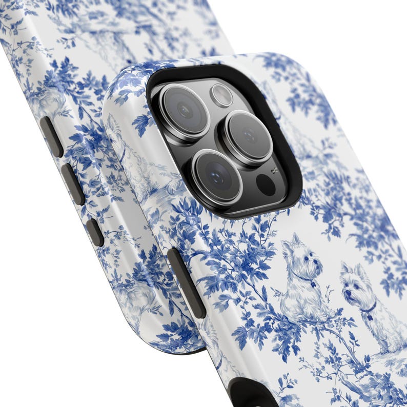 May include: A white phone case with a blue and white floral pattern and two white dogs. The case has a black camera bump with three lenses.