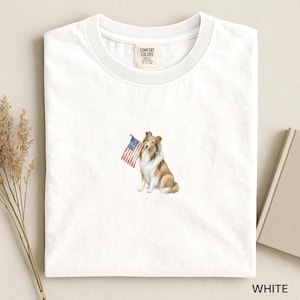 May include: A white crew neck t-shirt featuring a graphic of a brown and white Shetland Sheepdog sitting and holding a small American flag in its mouth. The shirt has a Comfort Colors brand label printed on the inside of the collar area.