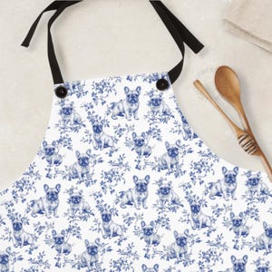 May include: White apron with a blue floral and French bulldog pattern. The apron has black ties and two black buttons. The design features multiple illustrations of French bulldogs and floral elements.