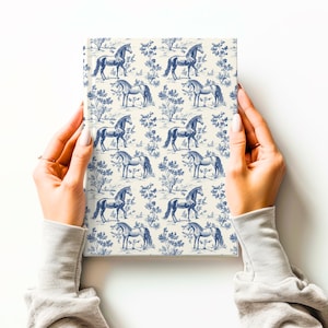 May include: A white notebook with a blue and white pattern of horses and trees. The notebook is being held by two hands.