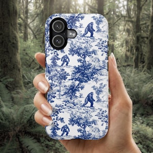 May include: A white and blue phone case featuring a toile design with Bigfoot figures in a forest setting. The case has a glossy finish and a camera cutout.