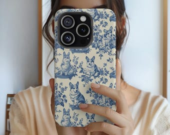 May include: A phone case featuring a blue and white toile pattern with images of corgi dogs and floral elements. The case has a cream-colored background and a glossy finish. The camera lenses are visible at the top.