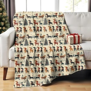 Puede incluir: Una manta crema con un patrón repetido de perros salchicha y árboles de Navidad. Los perros son de varios colores, incluyendo marrón, negro y beige. Un regalo envuelto con una cinta roja está en el sofá.