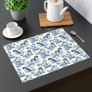 Könnte beinhalten: Ein rechteckiger Tischset mit cremefarbenem Hintergrund und einem blauen Toile-Muster mit Hunden und floralen Elementen. Das Tischset liegt auf einer dunklen Oberfläche, mit einer weißen Tasse und Untertasse, einer Teekanne und einer kleinen Pflanze.