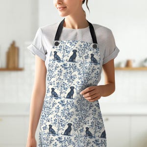 May include: An apron featuring a blue and white toile pattern with silhouettes of black dogs and floral elements. The apron has a black neck strap and button details. The design is repeated across the apron's surface.