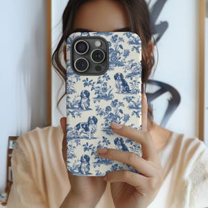 May include: A white phone case with a blue and white pattern of dogs and flowers. The case has a camera cutout and a clear back.