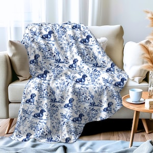 May include: A white and blue throw blanket with a repeating pattern of dachshunds and floral designs. The blanket is folded and draped over a white couch.