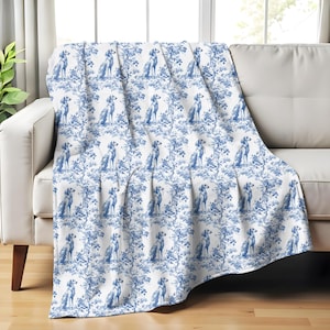 May include: A white throw blanket with a repeating blue pattern of dogs and foliage. The blanket is draped over a white sofa. The design is reminiscent of classic toile patterns, offering a decorative accent for home decor.