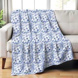 Könnte beinhalten: Eine blau-weiß gemusterte Decke, die über einem weißen Sofa drapiert ist. Die Decke zeigt ein sich wiederholendes Design von Hunden und Ästen. Das Farbschema erinnert an klassisches blau-weißes Porzellan.