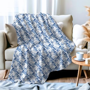 May include: A white fleece throw blanket with a repeating pattern of blue and white floral designs. The blanket is folded and draped over a white couch.