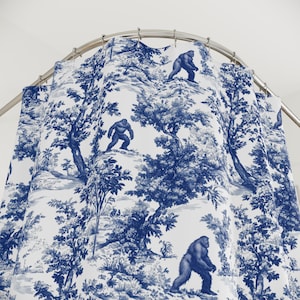May include: A blue and white shower curtain featuring a toile design with a forest scene and several depictions of a Bigfoot creature. The curtain is hanging from a curved metal rod.