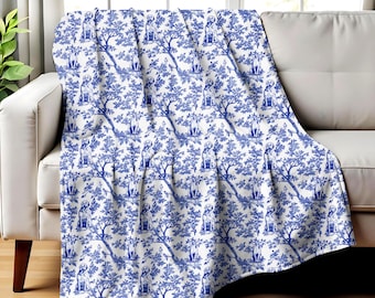 May include: A blue and white throw blanket draped over a white sofa. The blanket features a repeating pattern of dogs sitting under trees. The design is in a toile style, with a classic and elegant aesthetic.